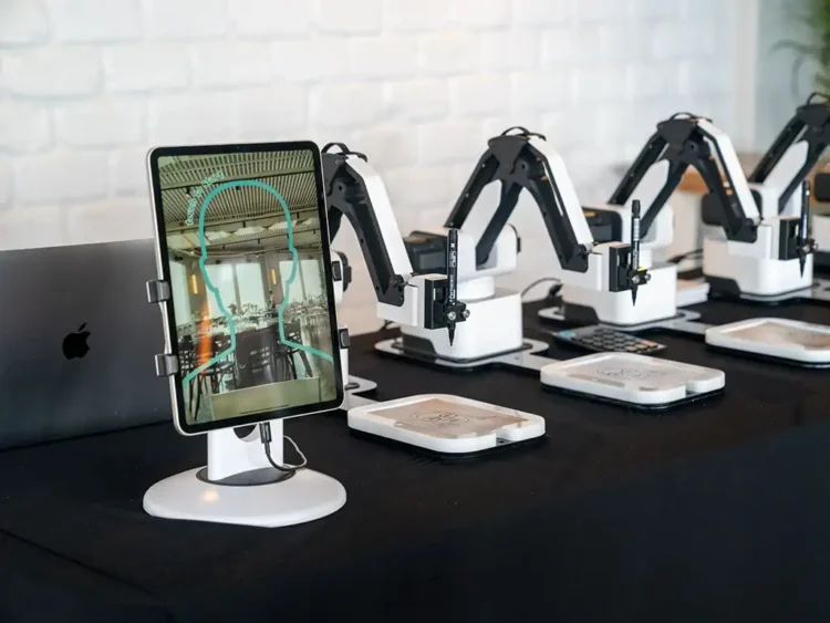 Porträtaufnahme von mehreren Sketch-Bot-Zeichnungsrobotern auf einem Tisch; im Vordergrund steht ein Tablet mit einer Linienzeichnung eines Kopfes auf dem Display, die Roboter zeichnen automatisch Porträts basierend auf digitaler Vorlage, Event- und Technik-Atmosphäre.